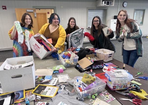 5 EcoReps smiling infront of the crafting table at the DIY day event. Crafting supplies donated and sourced from campus are on the table infront of the cohort members. Some members are holding up socks and  HSU t-shirts they upcycled